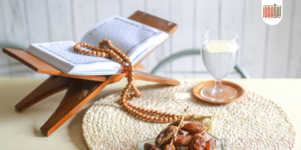 A glass of water on a prayer mat during Ramadan, with the Quran and dates in the background.